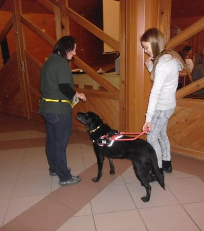 In the picture is a girl, holding on to the harness of a black labrador. They have just arrived back from their round. The trainer lady is standing in front of Dió, the guide dog.