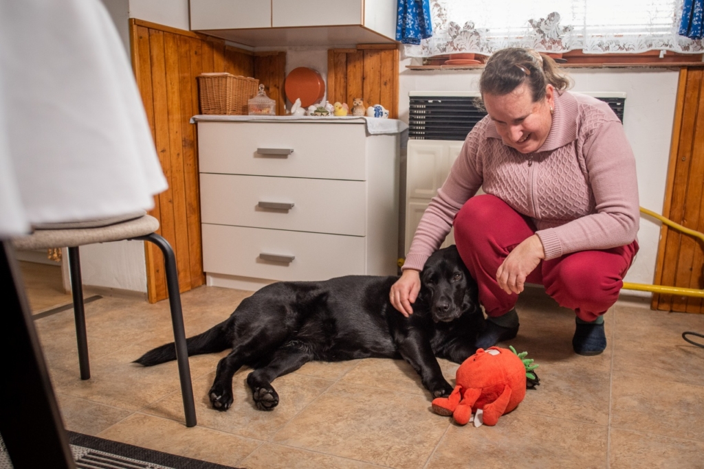 A képen Sáfrány Írisz látható, amint leguggolva simogatja nyugdíjas fekete labrador vakvezető kutyáját, Dollyt, aki mellette fekszik; Dolly előtt egy piros plüss játék látható.