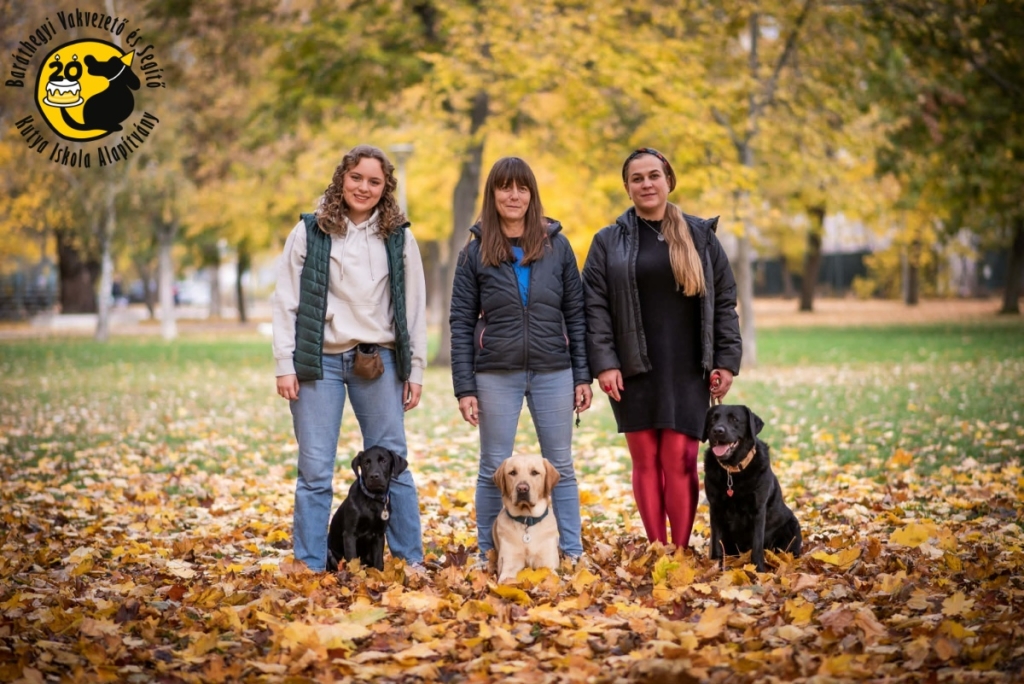 A képen egy őszi parkban három személy látható: középen Nyíriné Kovács Mária kiképző áll, bal oldalán egy kölyöknevelő önkéntes, jobb oldalán az egyik látássérült kliense; mindhármuk előtt egy labrador kutya ül.