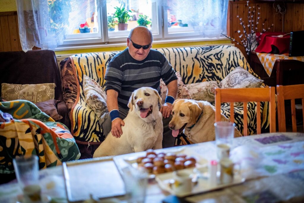 A képen Kiss Barna egy szobában, egy kanapén ül. Előtte két vakvezető kutyája látható: jobb oldalon Gyömbér, aki már nyugdíjas, bal oldalon pedig Tangó, az aktív vakvezető kutyája.