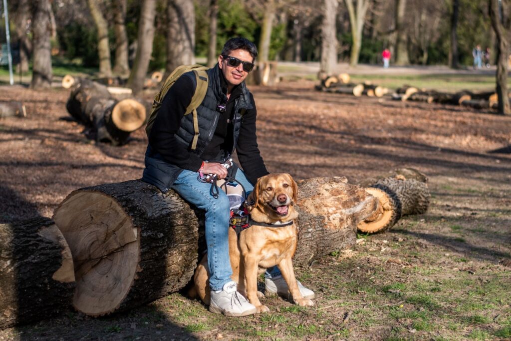 Bodnár Attila a parkban egy kivágott fatörzsön ül, előtte vakvezető kutyája, Parfé ül.