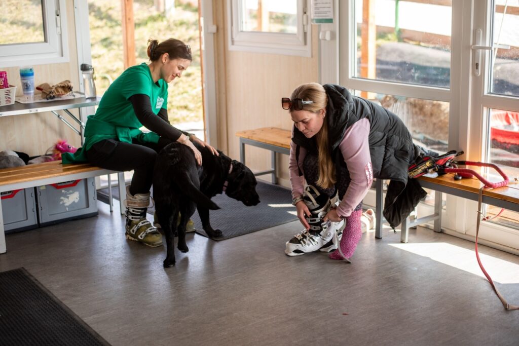 The visually impaired athlete Zsófi Lengyel sits on a wooden bench putting on ski boots, while her companion, a woman in a green shirt, pets the black Labrador guide dog in the background. They are in a bright, spacious building with wooden floors and windows.