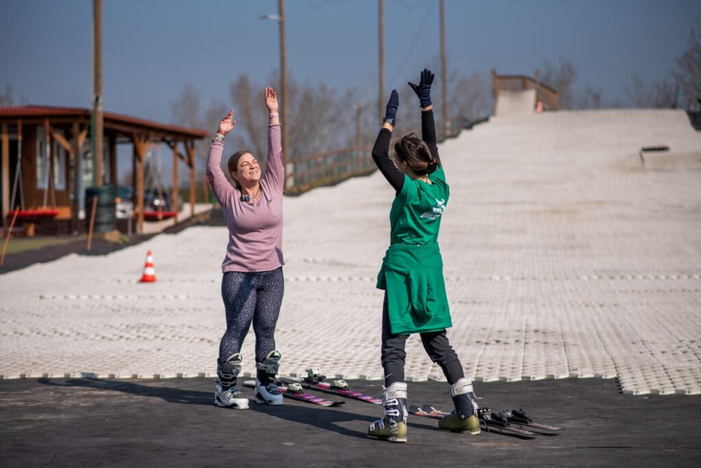 Zsófi Lengyel, the visually impaired skier, wearing a pink top, and her companion in a green shirt stand in ski boots at the bottom of the plastic slope, doing warm-up exercises with raised arms before skiing.