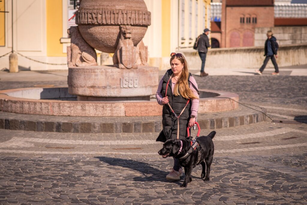 Zsófi Lengyel walks with her black Labrador guide dog, Mokka, in front of a fountain.