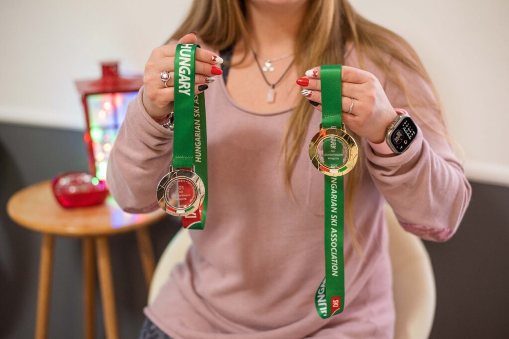 Zsófi Lengyel proudly shows the two medals she won in ski competitions. In her left hand is a silver medal, and in her right hand a gold medal, both hanging on green ribbons marked ‘HUNGARIAN SKI ASSOCIATION.’