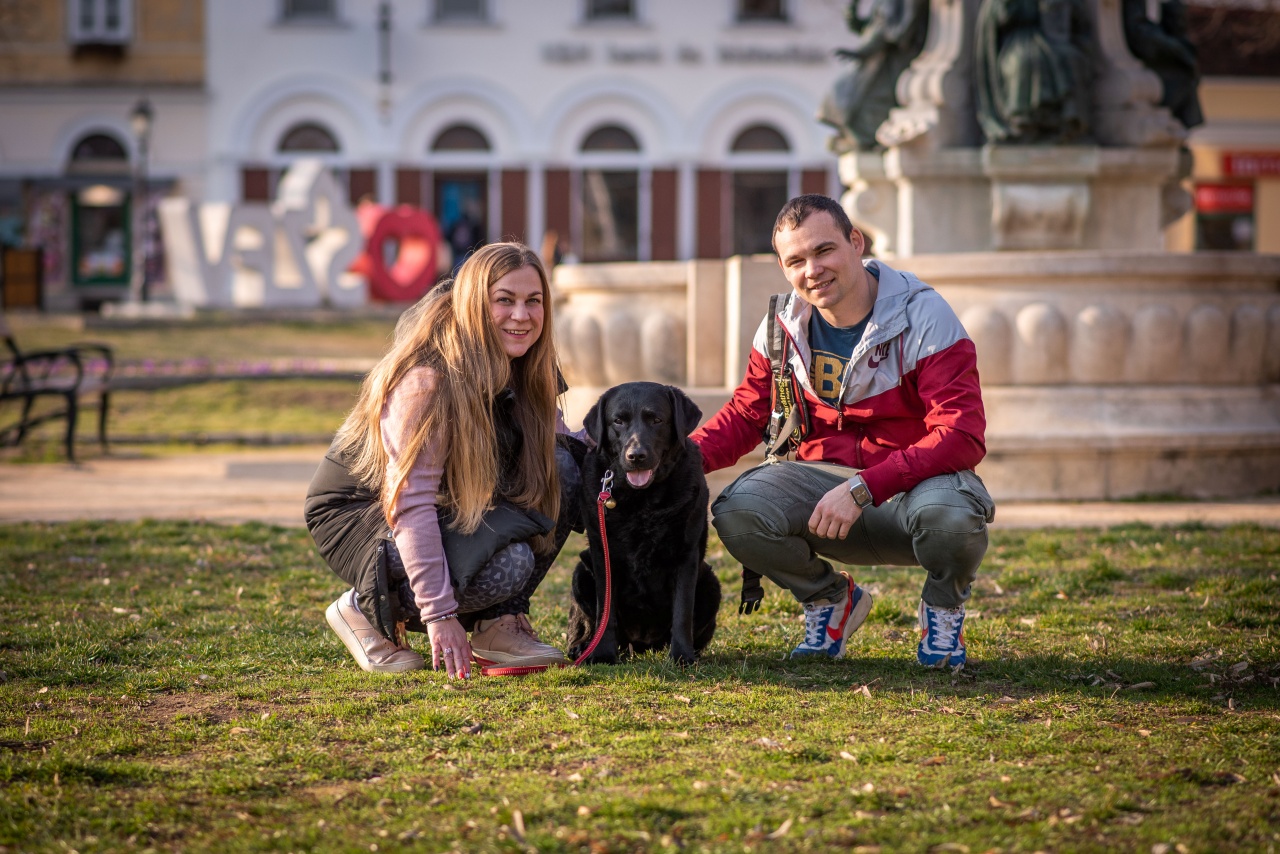 Lengyel Zsófi és párja, Tamás, egy parkban guggolnak a fűben, fekete vakvezető kutyájukkal, Mokkával. A háttérben egy szökőkút és városi épületek látszanak.