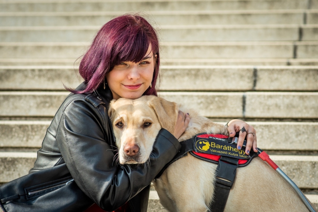 Flóra egy képcső alján ül és átöleli vakvezető kutyáját.