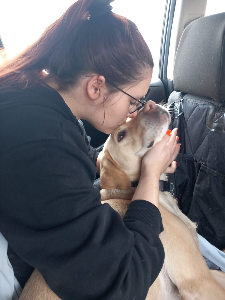 Egy autóban egy fiatal nő szeretetteljesen puszit nyom egy sárga labrador vakvezető kutya fejére, miközben az arcát a tenyerébe fogja.