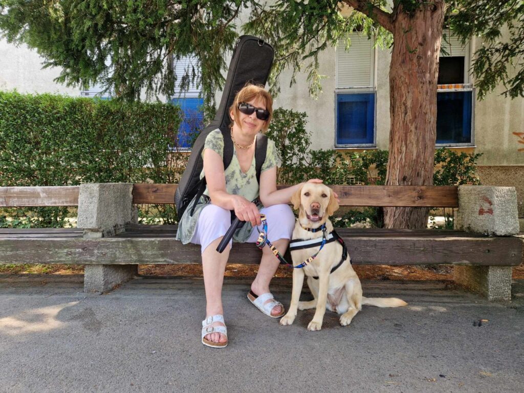 Egy napszemüveges nő egy parki padon ül, hátán gitártokkal, mellette pedig egy hámot viselő sárga labrador vakvezető kutya ül figyelmesen.