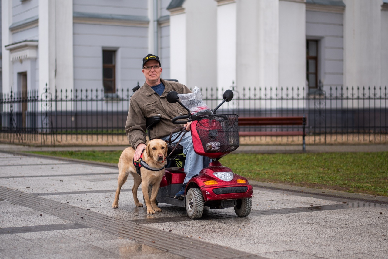A képen Göncző László mopedjével egy téren áll, mellette Frappé, a segítő kutya, aki mellette áll, miközben László a kezét a kutya hátára teszi.