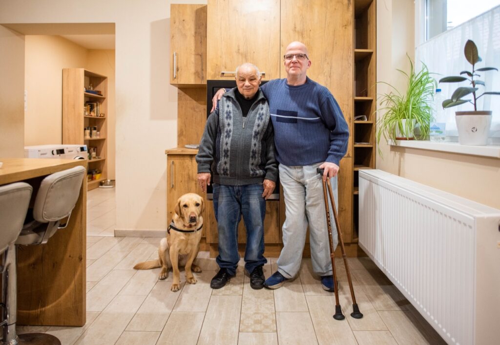 A képen Göncző László látható a Baptista Tevékeny Szeretet Misszió közösségi terében, mellette egyik lakótársa áll, akinek a vállát átöleli; mellettük ül Frappé, a segítő kutyája.