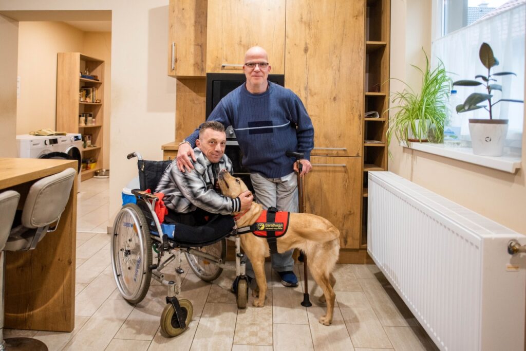 A képen Göncző László látható a Baptista Tevékeny Szeretet Misszió közösségi terében; mellette egy kerekesszékes lakótársa áll, akinek a vállát átöleli, előttük pedig Frappé, a segítő kutya áll, akit a kerekesszékes lakó szeretettel átölel.