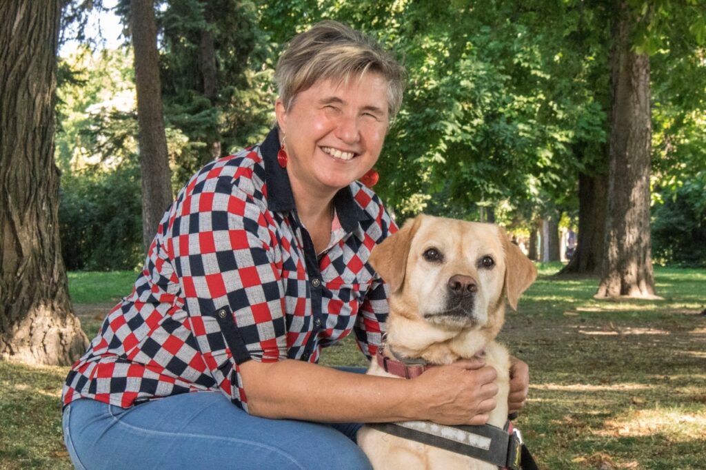 A képen Borsodi Erzsébet látássérült nő látható vakvezető kutyájával, Gesztenyével; a kutya ül, Erzsébet mellette guggol egy parkban, és szeretettel átöleli Gesztenyét.