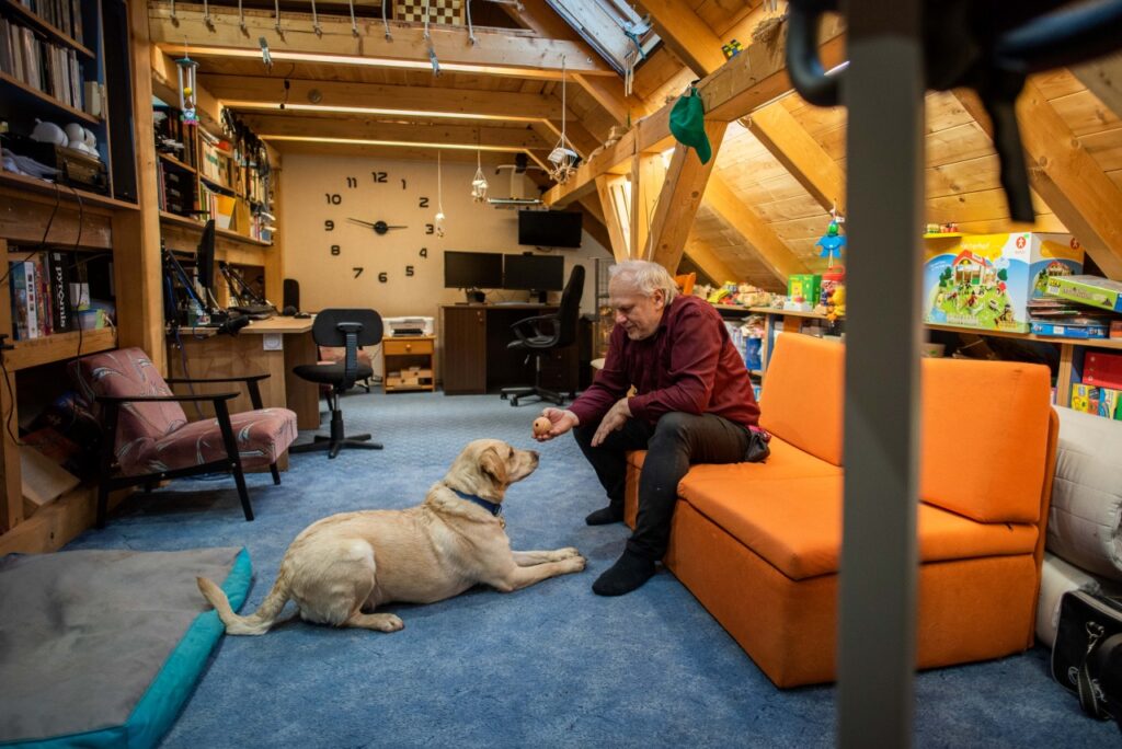 Bíró Miklós egy tetőtéri szobában egy narancssárga kanapén, és egy labdát mutat a vele szemben fekvő sárga labrador vakvezető kutyájának Atomnak. A háttérben íróasztal, számítógépek és egy nagy falióra látható.