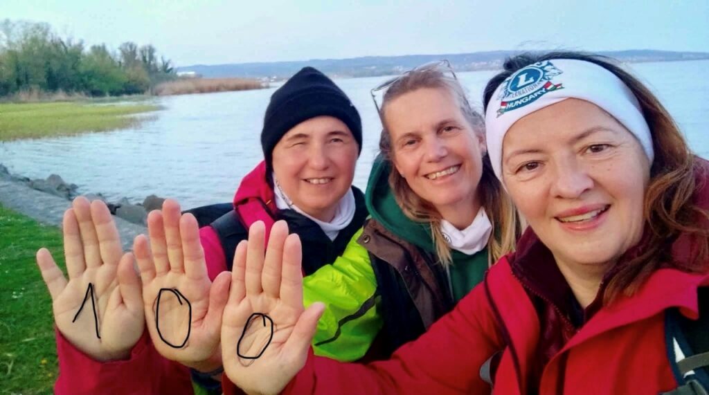 A képen három hölgy látható, akik egymás mellé helyezik a tenyerüket, rajtuk a „100” felirattal; jobb oldalon Borsodi Erzsébet, látássérült hölgy látható, mögöttük a Balaton.