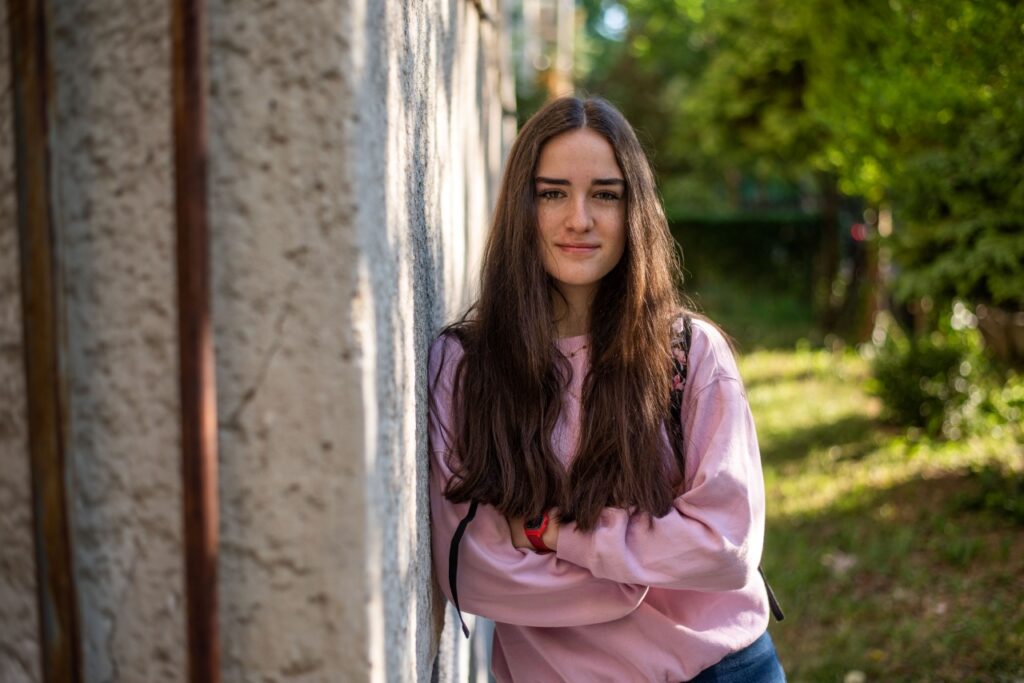 Hosszú barna hajú, szeplős fiatal nő mosolyog a kamerába, karba tett kézzel támaszkodik egy házfal mellett a kertben, pink pulóverben.