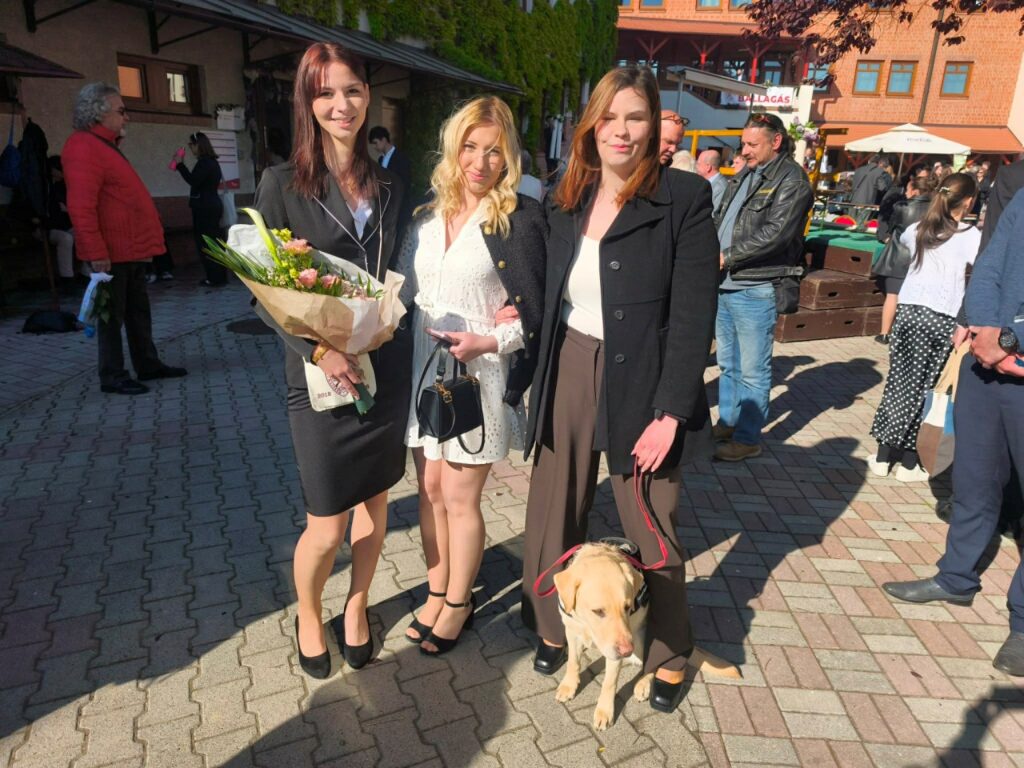 Három fiatal nő és egy sárga labrador vakvezető kutya áll egy térkövezett udvaron egy ünnepségen. Az egyik nő ballagási csokrot tart, a másiknál a vakvezető kutya póráza van.