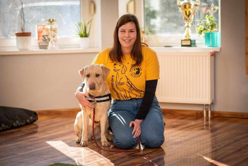 Zoé, az alapítvány látássérült önkéntese, sárga pólóban térdel a fényes padlón egy sárga labrador mellett. A kutya hámot visel, Zoé pedig átöleli a vállát és mosolyog.