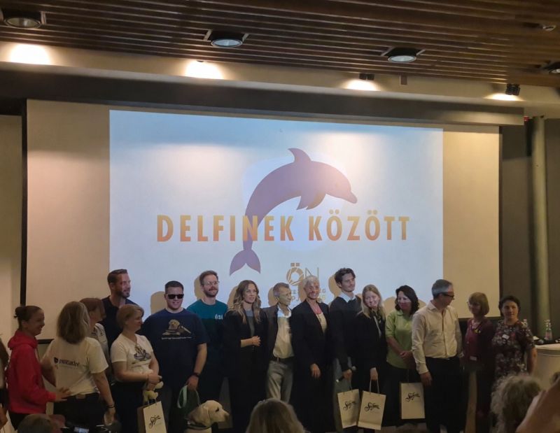 Group photo on stage: a larger group stands in front of a large projection screen showing a dolphin silhouette and the words "AMONG THE DOLPHINS."