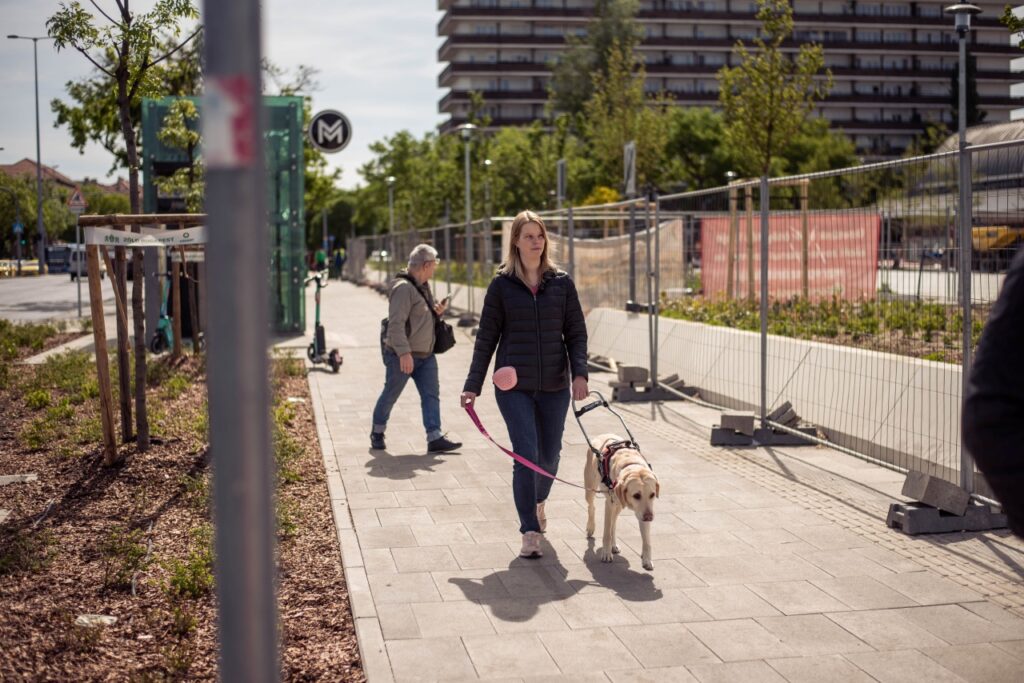 Réka fekete pufidzsekiben és farmerben sétál a sárga színű vakvezető kutyájával, Babérral egy napfényes városi járdán, építési terület és egy metrómegálló közelében