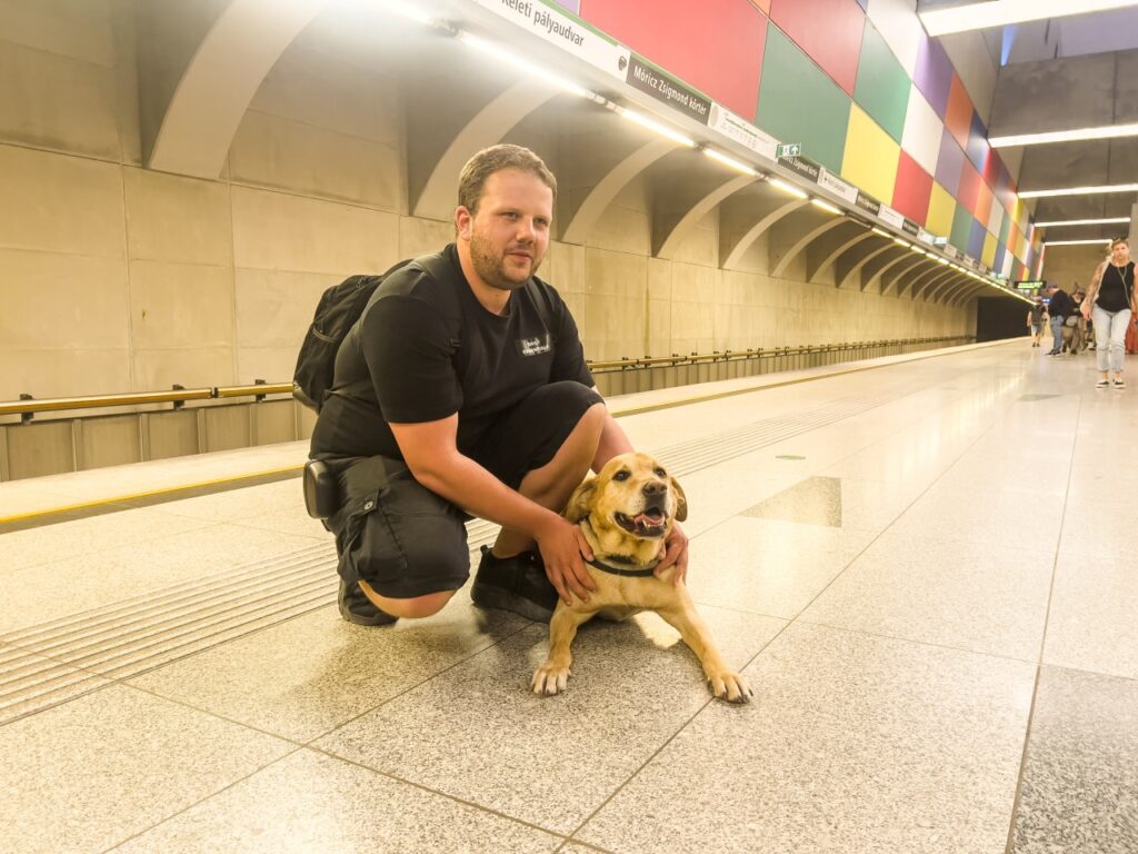 Orbán Roland egy metróállomás peronján guggol sárga színű vakvezető kutyája, Orion mellett, és mosolyogva tartja őt, miközben a kutya elégedetten fekszik a kőburkolaton.
