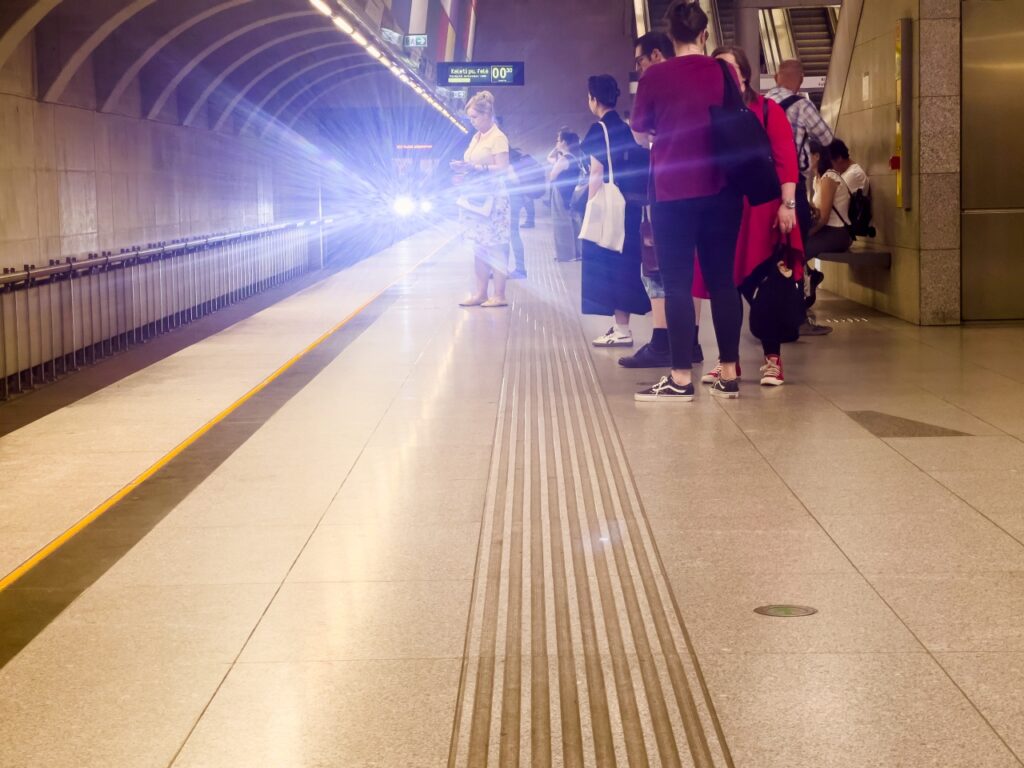 Utasok várakoznak egy metróállomás kivilágított peronján, miközben a távolból, az alagútból fénycsóvát szórva érkezik a metró szerelvény.