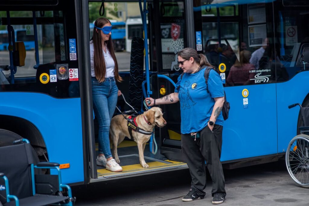 Egy bekötött szemű fiatal nő lép le egy kék BKK-busz nyitott ajtajából a vakvezető kutyájával, miközben egy kiképző segít nekik a BKK dolgozóinak tartott szemléletformáló tréningen.