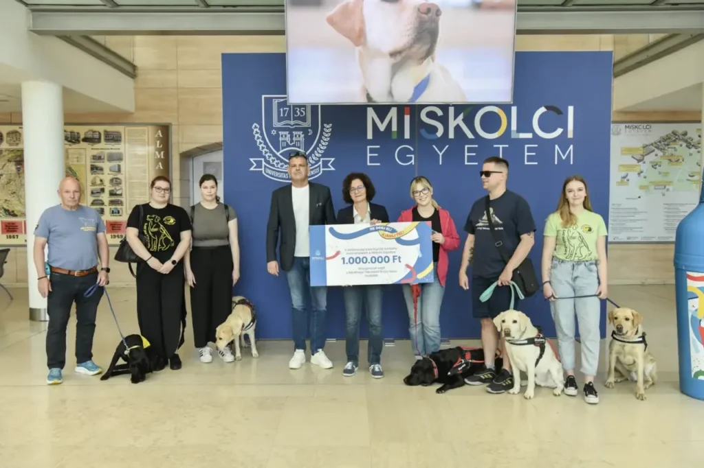 A Miskolci Egyetemen tartott adományátadáson nyolc ember és öt vakvezető kutya áll a „MISKOLCI EGYETEM” feliratú kék háttér előtt, miközben középen egy egymillió forintos adománygyűjtő csekket tartanak