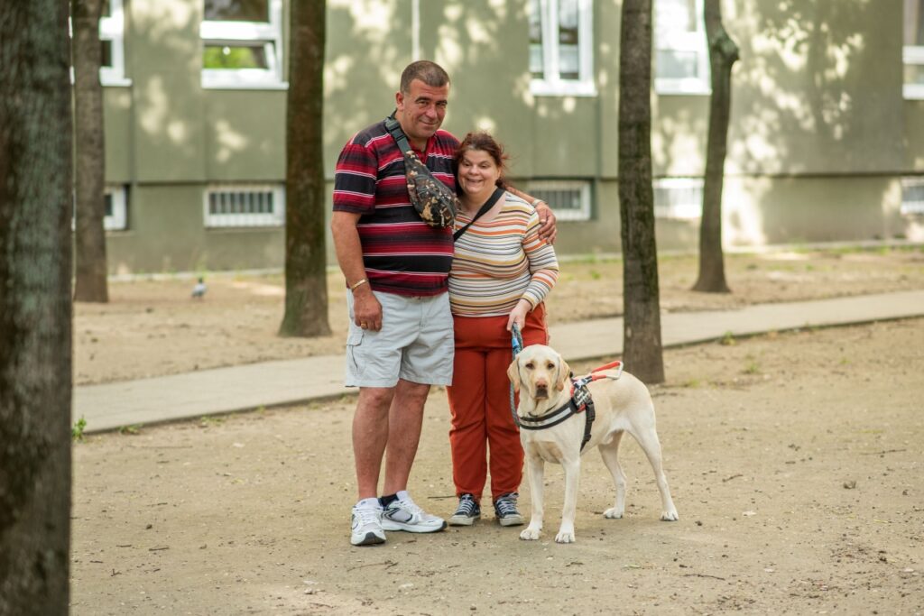 Kata és Zoli egymást átkarolva mosolyognak a kamerába a sárga színű vakvezető kutyájuk, Dióda mellett egy fás parkban.